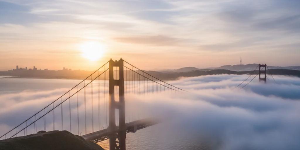 The Golden Gate Bridge rising out of low fog at sunrise, towers in International Orange against a pale sky